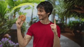 Young man in red shirt smiling holds glazed donut by face and points index finger in forest; enthusiasm. - Powered by Shutterstock - Get 15% off with code: PIKWIZARD15
