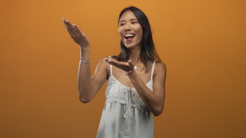 Thai woman wearing white sundress smiles and presents palms up with open hands while tilting head in studio; joyful play.