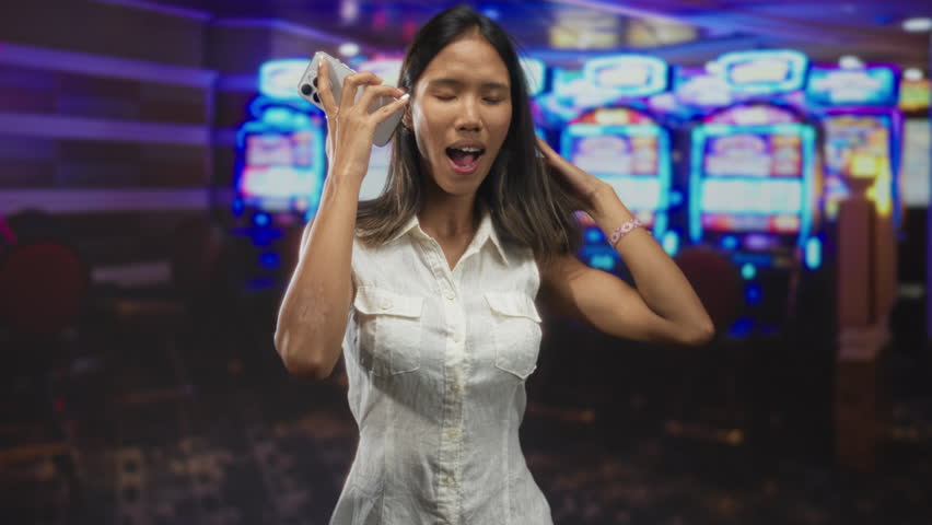 Woman holding smartphone to ear, talking and touching hair inside a casino building, sleeveless white dress and bracelet visible; amusement.