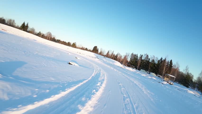 Dynamic FPV drone flight over a snow-covered racetrack at a winter motorsport venue. High-speed curves, drifting tracks, and clear skies during golden hour.