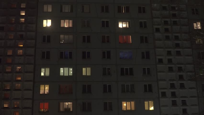 A nighttime view of a Soviet-era panel high-rise building. Many windows are illuminated with warm light, contrasting with the dark concrete facade. Light snow falls in this residential urban scene.