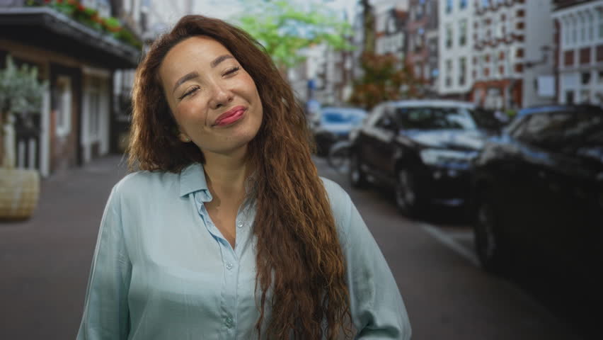 Woman smiling with visible face and long hair tilting head on city street, wearing light blue shirt; contentment daydream.
