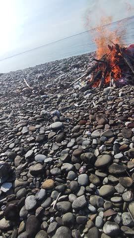 Namrole, Maluku Indonesia 12 December 2025.

A large bonfire crackles on a rocky beach as a person stands near the calm ocean under a blue sky. Ideal for themes of travel, summer, relaxation, and natu