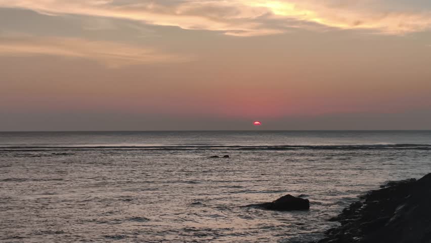Cinematic scenic sunset over the ocean with pink and orange clouds. Calm sea waves flowing near silhouette rocks during golden hour twilight. Peaceful and dramatic nature background.