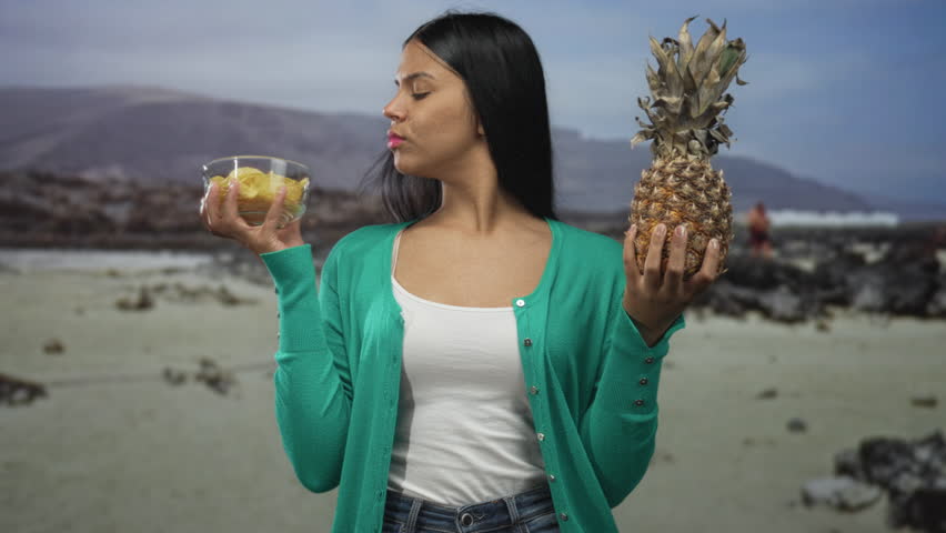 Woman wearing green cardigan uses both hands to hold bowl of chips and pineapple at seaside while squinting; indecision.