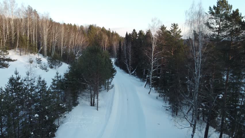 Aerial view of snowy winter landscape featuring forest, quiet road, falling snow, fir trees, and distant village

