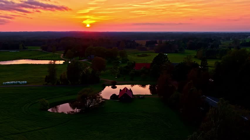 Gentle sunset colors spread across peaceful countryside fields. Time lapse with a reflective pond and scattered homes, creating a warm rural atmosphere captured from an elevated perspective.