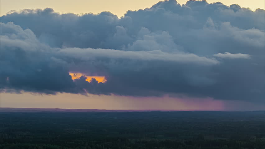 Dark storm clouds rolling over distant landscape with streaks of rain and a warm glow breaking through the cloud layer in a dramatic timelapse sky.