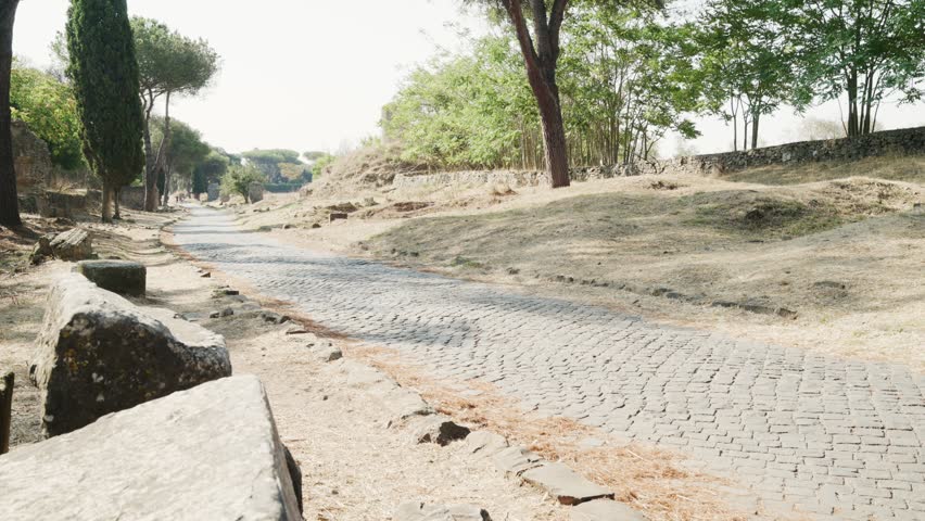 Young couple holding hands and walking along the historic Appian Way, an ancient cobblestone road in Rome surrounded by pine trees and archaeological ruins on a sunny day in Italy