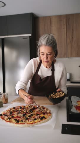 Older woman in a brown apron spreading fresh ingredients on a pizza base in a modern kitchen