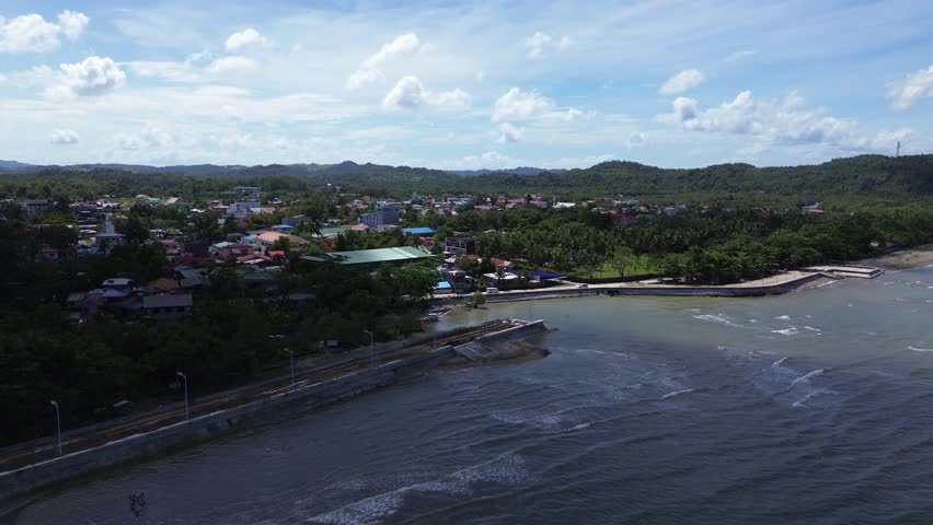 Aerial drone footage of a coastal promenade and concrete seawall in Odiongan City, Philippines, with ocean waves, sandy shoreline, city buildings and green hills under a sunny sky in Background.