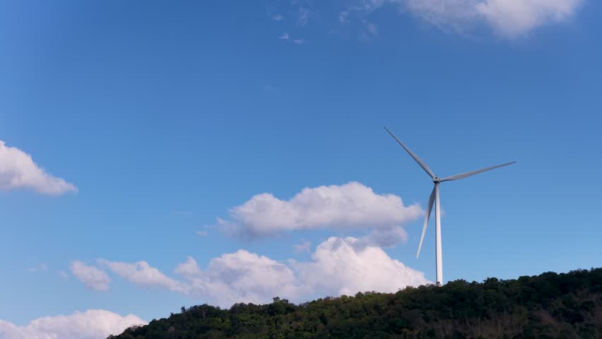 A static wide shot captures a wind turbine spinning against a bright blue cloudy sky