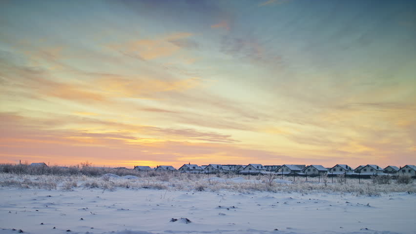Peaceful winter landscape with snow-covered ground and houses under a vibrant sunrise sky filled with pastel colors and clouds.