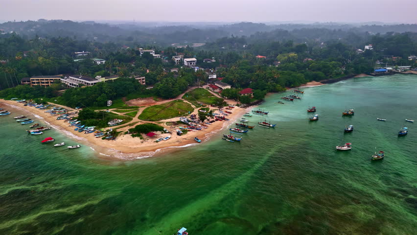 Drone aerial of Weligama fishing village on Sri Lanka’s south coast, with colorful boats lining a sandy peninsula and floating in a shallow turquoise bay.