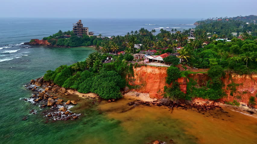 Drone view of Weligama’s red cliffs and rocky shoreline with palm trees, village houses and green water of the Indian Ocean on Sri Lanka’s south coast.
