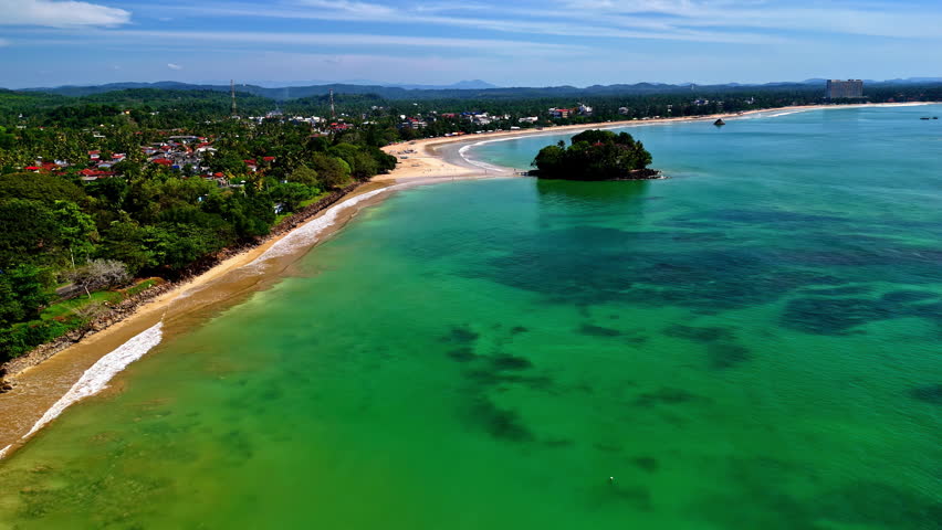 Aerial drone shot of Weligama Bay in southern Sri Lanka, showing a long curving sandy coastline, turquoise shallows with dark reef patches, and a small tree-covered island near the beach.
