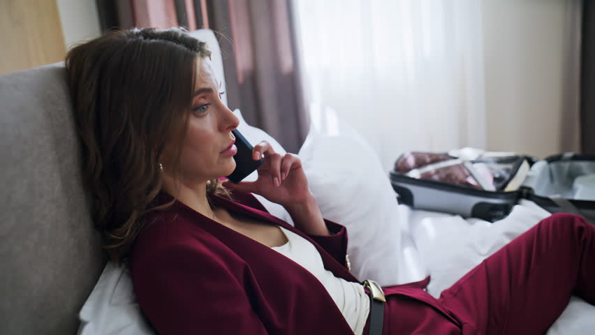 Hotel guest calling on bed discussing work closeup. Serious business woman wearing elegant suit talking cellphone lying in comfortable white bedding. Successful lady arranging meeting in business trip