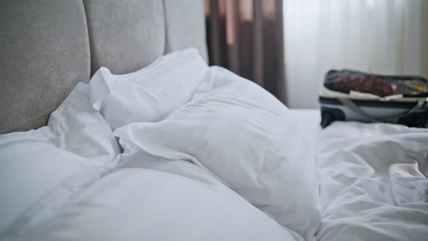 Hotel guest falling bed near travel suitcase closeup. Smiling beautiful woman lying on cozy white bedding enjoy luxury room interior. Carefree lady stretching relaxing in light bedroom. Travel concept