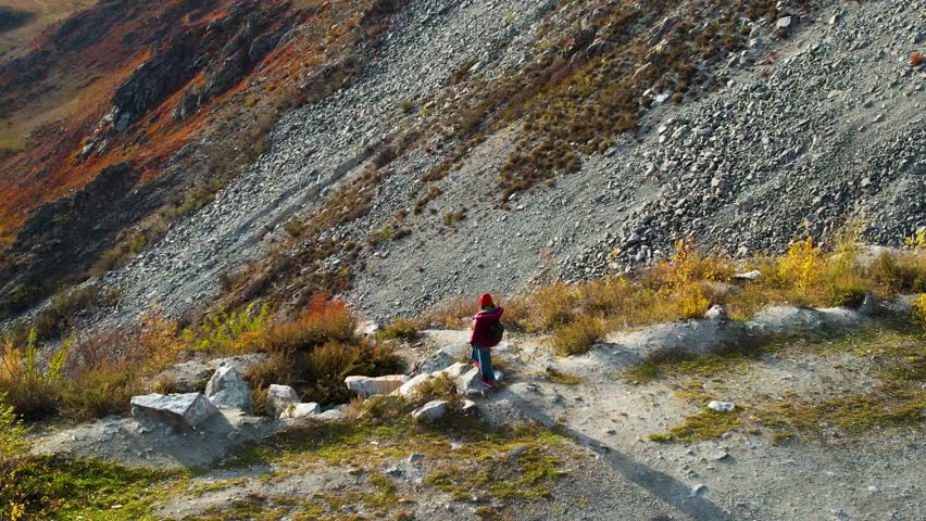 Hiker In Autumn Mountain Valley Pauses Beside Rocky Slope And Golden Foliage, Aerial View Reveals Expansive Rugged Terrain, Solitary Figure In Red Jacket With Backpack Scans Vista, Long Shadow
