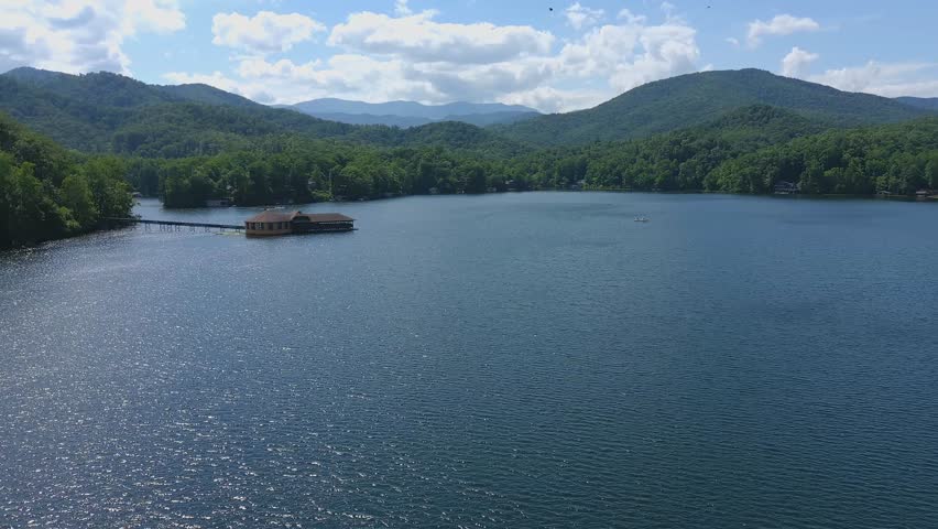 Panoramic view of a tranquil lake in Great Smoky Mountains National Park, surrounded by lush forested hills and distant peaks, with a wooden cabin and pier extending into calm reflective water under a partly cloudy sky.
