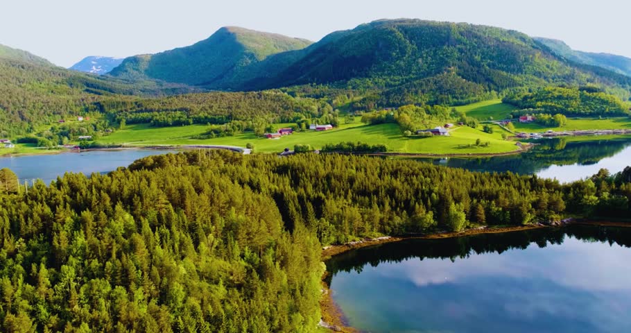 Beautiful aerial view of a lush green countryside with rolling hills, dense forest, calm river and scattered rural houses. Scenic Nordic landscape with mountains, farmland and reflective water under clear daylight. Peaceful nature background, rural life, sustainable environment and travel destination concept.