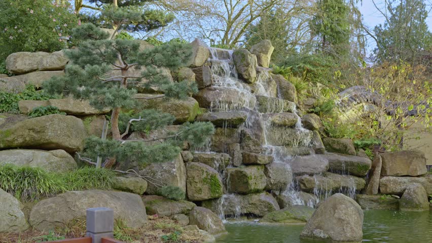 A serene Chinese garden in Parc de Maurepas, featuring a rock waterfall and a beautifully shaped bonsai tree. Ideal for travel, cultural, and relaxation concepts, Rennes in France