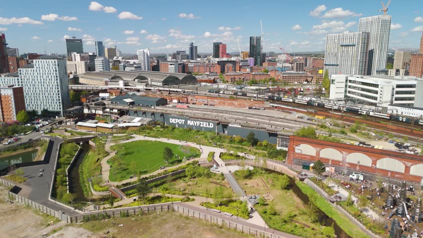 Descending circling drone shot of Manchester city centre landscape with trains pulling into train station. Blue sky with fluffy clouds and green park below