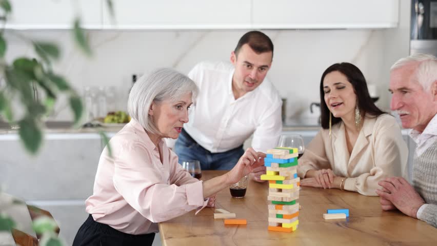 Group of mature spouses and middle-aged male visitor with girlfriend is playing board game. Senior married couple build tower with wooden blocks bricks with younger spouses