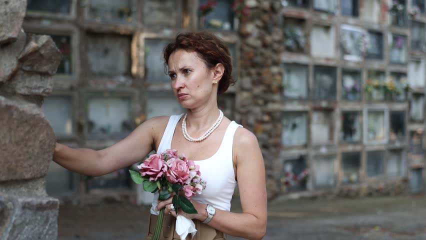 Sad young woman in white croptop with bouquet of flowers and handkerchief in hands, stands in cemetery. Visiting grave of deceased relative, mourning dead loved one. Appeal to God, prayer for dead