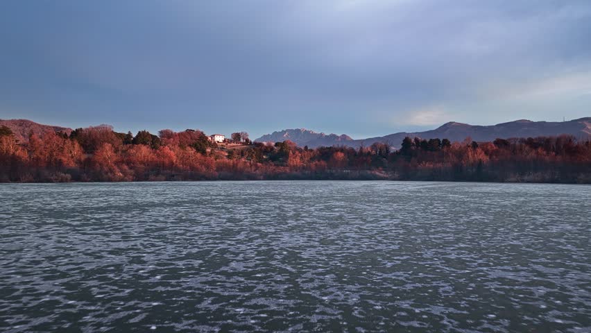 Aerial drone shot flying low forward over a frozen lake at sunrise, revealing winter forest and distant mountains, calm and atmospheric early morning landscape.