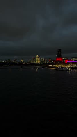 Illuminated london skyline glowing at night over the dark, rippling water of the river thames, under a cloudy, dramatic evening sky