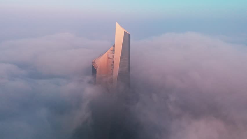 Cinematic drone aerial of Al Hamra Tower in Kuwait City emerging through thick fog and low clouds at sunrise, glowing with warm light above a soft blue sky.