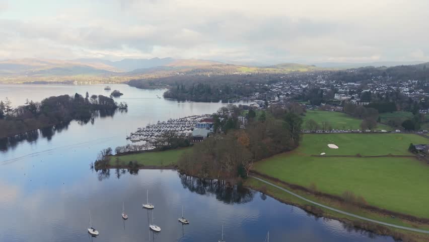 A left pan over Lake Windermere with land to the right and open water below. The drone passes toward a small marina as sunlight breaks through clouds, illuminating distant mountain peaks.