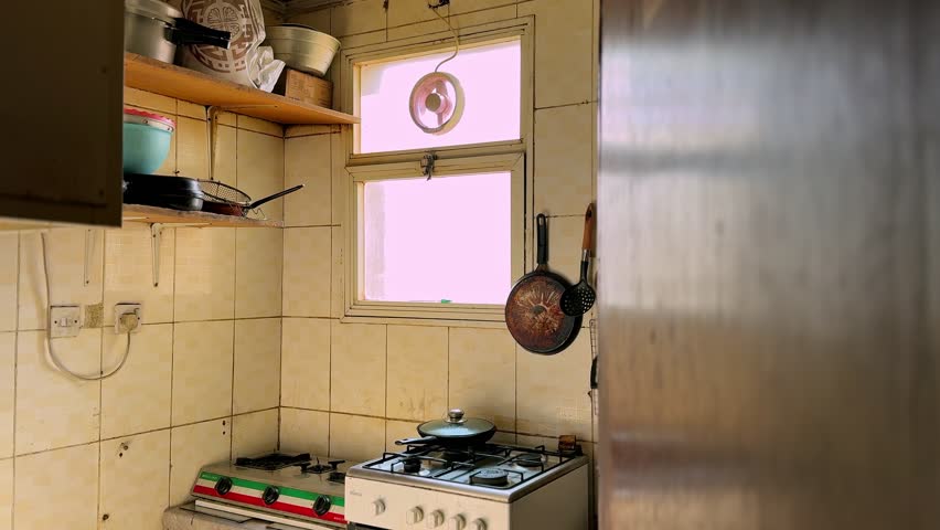 Static shot of rustic small kitchen with tiled walls, gas stove, hanging pans and utensils, open shelves, and bright window light in a lived-in home.