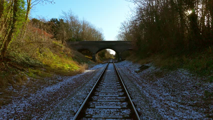 Drone flying above stone bridge crossing railway line through winter landscape. Aerial camera gliding over brick overpass spanning rail corridor among trees and frosty ground. Overhead travel scene