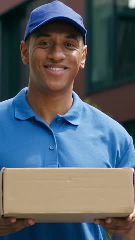 Happy smiling African American delivery man standing outdoors in city male courier holding package cardboard shopping order service looking at camera shipment joy postman professional worker handling