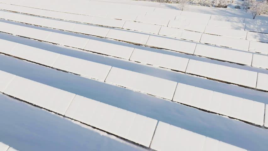 Aerial tracking shot in front of snow covered solar panels, sunny winter morning
