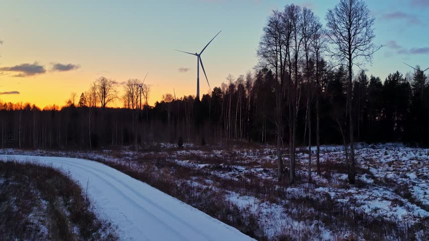 Snow-covered field and forest with wind turbine at sunset under colorful winter sky