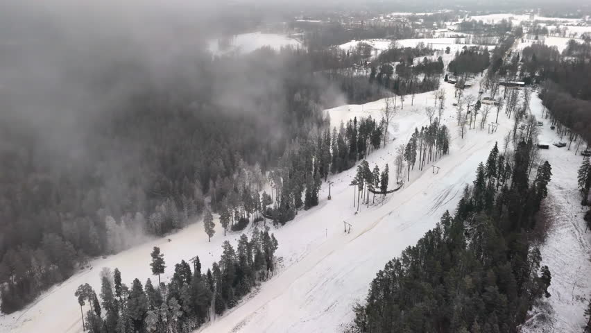 Clouds rise off top of forest in snowy slope corridor cutting through forested winter terrain, aerial pullback