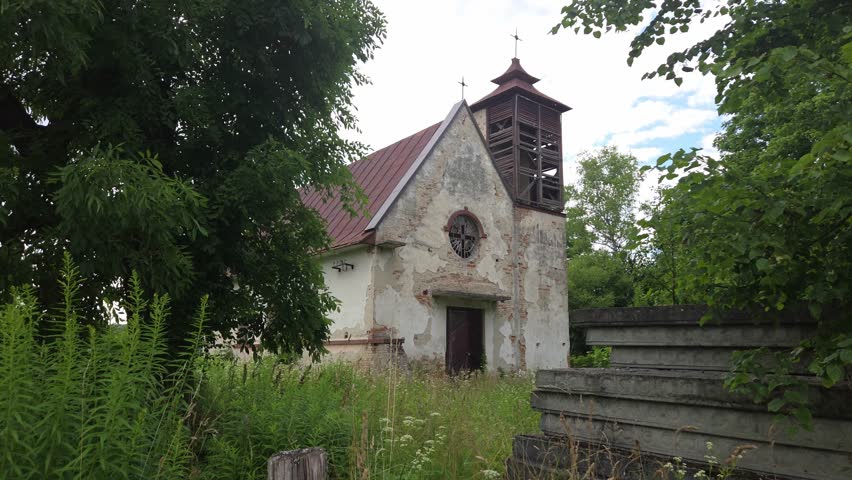 Weathered stone church with a wooden bell tower and red roof stands abandoned among tall green grass and lush trees under a cloudy summer sky, showcasing its historical architecture