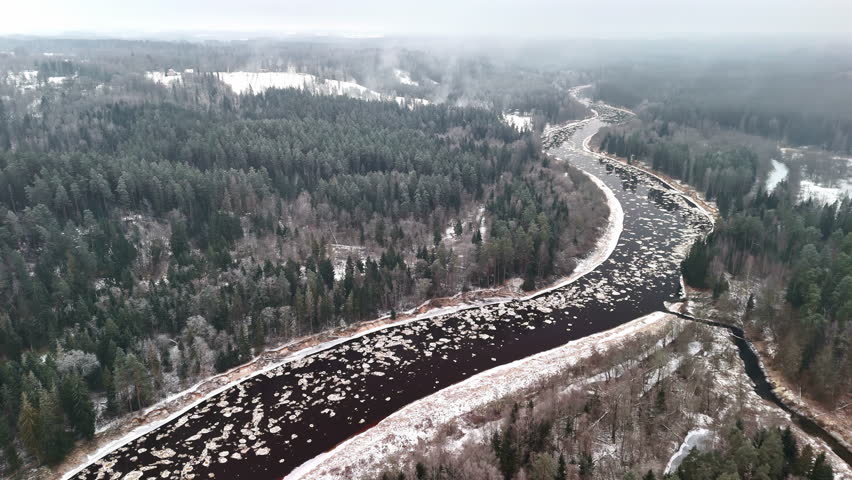 Aerial view of a winding winter river flowing through dense snowy forest, with drifting ice patches and misty cold air above the landscape.