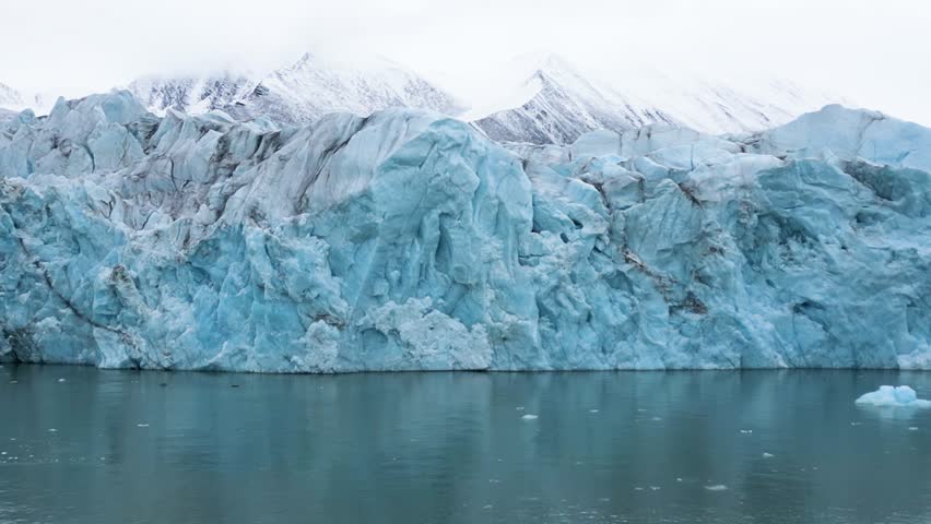 Massive blue tidal glacier front moving along cold arctic waters with small boat and rugged snowy mountains above