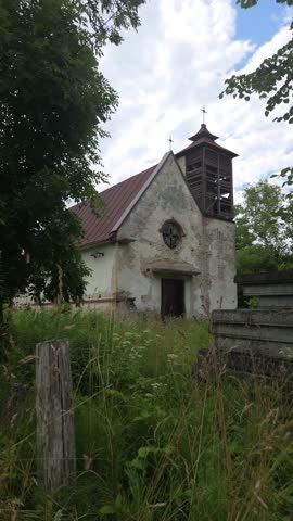 Weathered stone church with a wooden bell tower and red roof stands abandoned among tall green grass and lush trees under a cloudy summer sky, showcasing its historical architecture