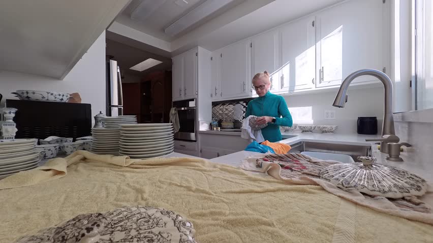 A mature woman washes antique heirloom china bowls and plates in a bright kitchen. She carefully cleans each piece by hand.
