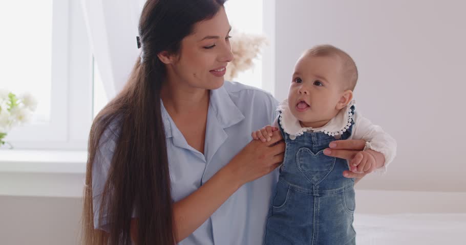 Happy young family of cheerful mother and little cute baby sitting together on bed in bedroom at home. Woman parent having fun with child. Family leisure, love and care concept. 4k video.