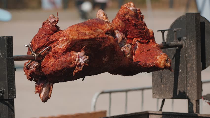 A whole pig roasts on a horizontal spit, revealing its crisp, seasoned skin. Grilled with glowing coals underneath. Traditional cooking method