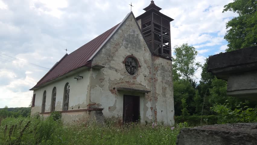 Abandoned stone church with a wooden bell tower and red roof stands among tall grass and lush trees under a cloudy summer sky, showcasing its weathered and historical architecture