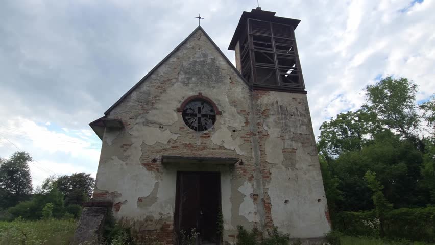 Weathered stone church with a wooden bell tower and red roof stands abandoned among tall green grass and lush trees under a cloudy summer sky, showcasing its historical architecture