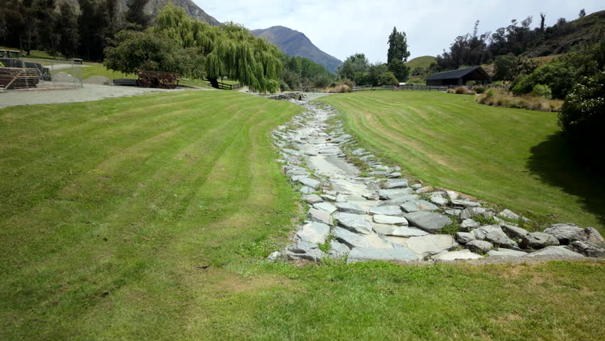 Stone Lined Creek Bed Through Green Park Lawn With Mountains