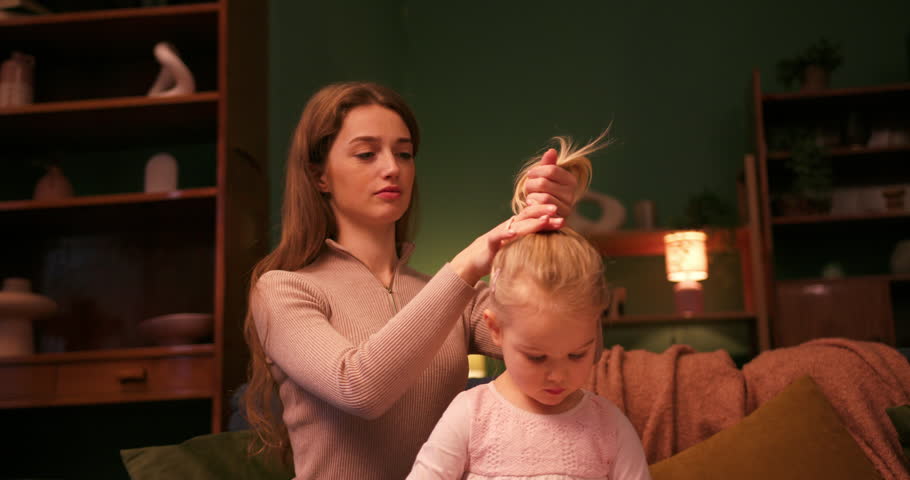 Loving mother styling her little daughter's hair into a ponytail while sitting on the sofa
